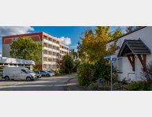 Mehrstöckiges Wohngebäude mit Balkonen im Hintergrund, umgeben von Bäumen in herbstlicher Färbung, sowie ein einstöckiges Gebäude mit Schrägdach im Vordergrund; ein Straßenschild mit den Namen "Wendenstraße" und "Kietz" ist sichtbar, daneben geparkte Autos und ein Transporter.