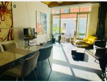 Wohnzimmer mit modernem Design, ausgestattet mit einem großen Glastisch, gepolsterten Stühlen, einer gelben Liege und einer Glasfront, die auf einen Balkon mit Pflanzen und Blick auf gegenüberliegende Gebäude führt. Die Wände sind mit abstrakten Gemälden dekoriert.