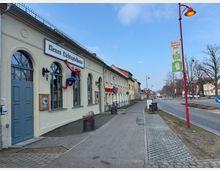 Straßenszene mit einem Gebäude im Vordergrund, auf dem „Neues Lichtspielhaus“ steht und ein an die Wand modellierter Spider-Man angebracht ist. Rechts neben dem Bürgersteig befinden sich eine Straße, rote Laternenmasten und Werbeschilder, umrahmt von Gebäuden und kahlen Bäumen.