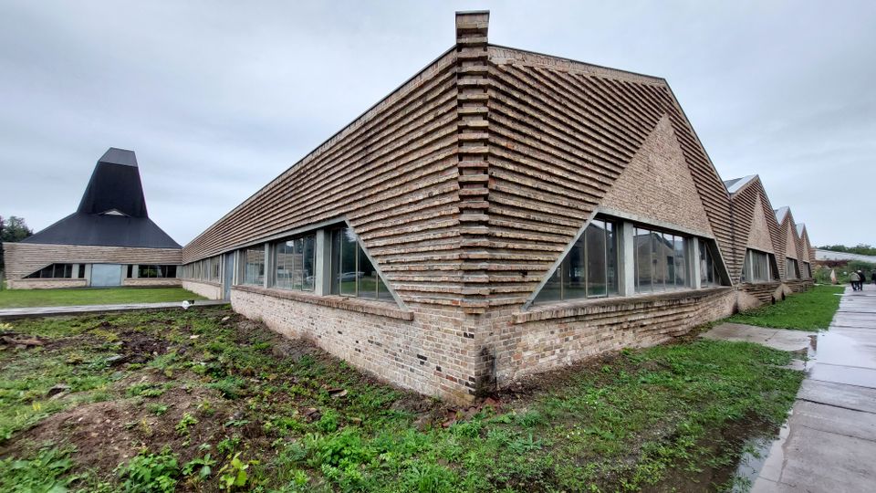 Ein modernes Gebäude mit markanter Klinkerfassade und gezacktem Dachdesign, ergänzt durch große Glasfenster. Im Hintergrund ist ein weiteres Gebäude mit einer schwarzen, pyramidenförmigen Dachkonstruktion sichtbar, umgeben von einer Grünfläche und Gehwegen.
