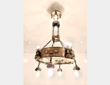 Großer, an Ketten aufgehängter Kronleuchter aus Metall im Industrie-/Steampunk-Stil mit genieteten Ringen und mehreren freiliegenden Glühbirnen. Hängt an einer hellen, weißen Decke mit weiteren kleinen Deckenleuchten im Hintergrund.