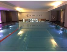 Schwimmbad in einem modernen Fitnessstudio mit rechteckigem Becken, beleuchteter Wasseroberfläche und einer Trennleine. Im Hintergrund steht "Holmes Place" an einer weißen Wand, umgeben von grauen Wänden und dezentem Licht.
