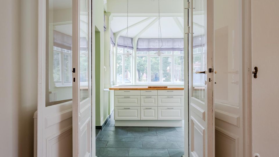 Blick durch geöffnete weiße Flügeltüren in eine helle Küche oder Veranda mit großflächigen Fenstern und gewölbtem Deckenbereich. Vor den Fenstern steht ein weißer Unterschrank mit Holzarbeitsplatte auf dunklem Steinfliesenboden.