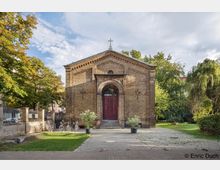 Backsteinkirche mit rechteckiger Fassade, einem zentralen Rundbogenportal mit roten Doppeltüren und einem Kreuz an der Spitze. Die Kirche ist von Bäumen umgeben, mit einem gepflasterten Vorplatz und einigen Blumentöpfen am Eingang.