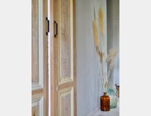 Eine Nahaufnahme eines Innenraums mit einer Holztür in natürlichem, abgenutztem Look auf der linken Seite. Rechts steht eine braune Glasvase mit getrocknetem Pampasgras vor einer hellgrauen Wand.