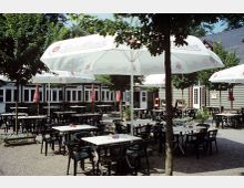 Ein Biergarten mit zahlreichen Tischen und Plastikstühlen auf einem Kiesboden. Große weiße Sonnenschirme spenden Schatten, und im Hintergrund ist ein Gebäude mit Fenstern und Türen sichtbar, umgeben von Bäumen.