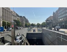 Blick auf den U-Bahn-Eingang "Unter den Linden" in einer breiten Stadtstraße, flankiert von historischen Gebäuden mit großen Fenstern. Im Vordergrund befinden sich Rolltreppen, Fahrräder sind abgestellt, und im Hintergrund sind Bäume und weitere Architektur zu sehen.