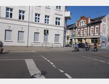 Straßenkreuzung in einer Stadt mit einem weißen Wohngebäude im Hintergrund, daneben ein Geschäftshaus aus roten Backsteinen. Drei Motorradfahrer im Vordergrund fahren bei grüner Ampel über die Kreuzung.