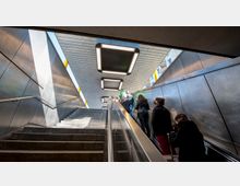 Eine Treppe mit Metallgeländern, die aus einer U-Bahn-Station nach oben ins Freie führt. Die Umgebung besteht aus metallischen Wänden und einer Decke mit rechteckigen Deckenlampen. Mehrere Personen gehen die Treppe hinauf.