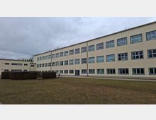 Beiger, mehrstöckiger Schul- oder Verwaltungsbau mit langen Reihen rechteckiger Fensterfronten und flachem Dach. Davor liegt eine große Rasenfläche mit niedrigen Hecken und einem Gehweg unter bewölktem Himmel.