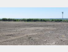 Weite, unbebaute Fläche mit braunem, trockenem Boden und vereinzeltem Bewuchs, im Hintergrund niedrige Erdaufschüttungen mit Gras und ein Metall-Funkturm. Der Himmel ist klar und blau.