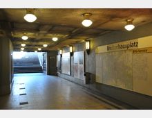 U-Bahn-Eingang in einem unterirdischen Gang bei der Station "Breitenbachplatz" in Berlin; an den Wänden befinden sich beleuchtete Hinweisschilder, Stadtpläne und Informationen. Im Hintergrund sind Treppen, die ins Freie führen, sowie mehrere Deckenlampen mit gelbem Licht sichtbar.