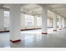 Großer, leerer Industrie- oder Loftinnenraum mit weißen Stützpfeilern, freiliegenden Deckenbalken und weitem Betonboden. Entlang der Außenwand befinden sich große, mehrteilige Sprossenfenster, die viel Tageslicht in den Raum lassen.