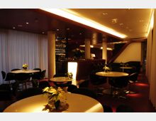 Elegantes Innenraumdesign einer Lounge oder Bar mit gedämpfter Beleuchtung, runden Tischen, schwarzen Stühlen und dezenten Dekorationen wie Weihnachtssternen. Im Hintergrund befindet sich eine beleuchtete Theke, flankiert von weißen Säulen und Vorhängen an den Fenstern.