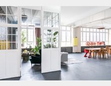 Großzügiger, heller Innenraum eines Lofts mit weiß gestrichenen Wänden, großen Fenstern und modernen Möbeln. Links ein separater Raum mit Glastüren, üppigen Pflanzen und einem Sessel, rechts ein Holztisch mit Stühlen und ein leuchtendes "Grand Hotel"-Schild an der Wand.