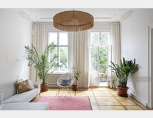 Wohnzimmer mit hohen Fenstern, durch die Tageslicht einfällt, weißen Wänden mit Stuckverzierungen und einem Parkettboden. Der Raum ist mit einem großen Sofa, Zimmerpflanzen, einem runden Korbsessel und einem rosafarbenen Teppich eingerichtet; ein geflochtener Hängelampenschirm hängt in der Mitte.