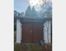 Freistehende, kleine Garage mit weiß verputzten Seitenwänden und großem, zweiflügeligem Holztor in Dunkelbraun unter einem flachen Dachüberstand. Davor unbewachsener Boden mit Grasresten; im Hintergrund Bäume und Sträucher, rechts ein kahler kleiner Baum und ein Verkehrsschild am Rand.
