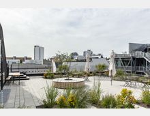 Dachterrasse mit Sitzbereichen, Tischen, Sonnenschirmen und Pflanzenbeeten, umgeben von modernen Gebäuden und einer urbanen Skyline im Hintergrund. Die Terrasse ist mit hellen Steinplatten ausgelegt und verfügt über eine zentrale, bepflanzte Sitzgelegenheit.