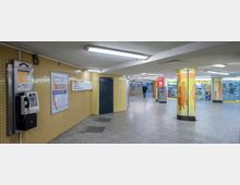 Unterirdischer Durchgang mit gelb gefliesten Wänden und grauem Boden. An den Wänden befinden sich ein Münztelefon, Werbeschilder und ein Orientierungsschild, während ein paar kleine Geschäfte sowie ein Geldautomat im Hintergrund sichtbar sind.