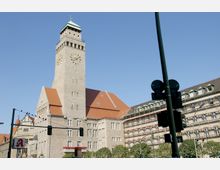Historisches Gebäude mit sandfarbenen Steinwänden und rotem Ziegeldach, dominiert von einem hohen Turm mit einer Uhr und grünem Kupferdach; im Vordergrund eine Straßenkreuzung mit Ampeln.
