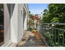 Ein schmaler Balkon mit Holzboden und Metallgeländer, geschmückt mit grünen und roten Pflanzen in Töpfen. Rechts steht eine bronzefarbene Metallbank, im Hintergrund sind Nachbargebäude und Bäume zu sehen.