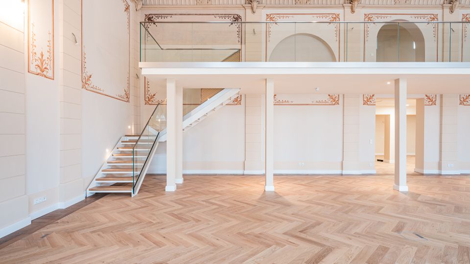 Heller, hoher Innenraum mit Fischgrät-Parkettboden, weißen Wänden mit dekorativen Stuck- und Ornamentrahmen sowie einer eingezogenen Galerieebene mit Glasgeländer. Links führt eine moderne Treppe mit Glasbrüstung zur oberen Ebene; im Hintergrund sind bogenförmige Durchgänge und Stützen zu sehen.
