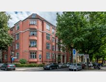 Eckgebäude im Wohnviertel mit rosa Fassade und mehreren Balkonen, umgeben von Bäumen an einer straßenkreuzenden Ecke. Im Vordergrund sind parkende Autos, Verkehrsschilder und Gehwege erkennbar.