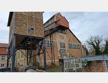 Backstein-Gebäude eines ehemaligen Industriekomplexes mit großen Fenstern, schrägem Dach und außen angebrachten Metalltreppen. Im Vordergrund befinden sich Metallkonstruktionen und Gerüste, im Hintergrund kahle Bäume und ein bewölkter Himmel.