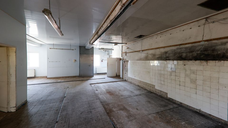 Leerer, heruntergekommener Industrie- oder Küchenraum mit gefliestem Boden, weißen Wandfliesen und deutlichen Ruß- und Schmutzspuren an der rechten Wand. An der Decke hängen lange Leuchtstofflampen und es verlaufen offene Leitungen; im Hintergrund sind Fenster und eine Tür zu sehen.