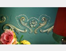 Wand mit grüner Oberfläche und goldenen, floralen Verzierungen im Barockstil; im Vordergrund unscharfe Blüten in Rosa und Gelb sowie ein roter Lampenschirm rechts.