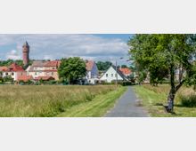 Ländliche Siedlung mit Einfamilienhäusern im Hintergrund, einem roten Wasserturm und einem schmalen gepflasterten Weg, der durch eine grüne Wiese führt; am Wegesrand steht eine Holzbank unter einem Baum.