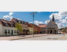 Straßenszene in einer Kleinstadt mit historischen Gebäuden, darunter ein Rathaus mit Turm und Spitzdach, sowie mehrfarbigen Wohnhäusern mit roten Ziegeldächern. Im Vordergrund befindet sich eine breite Straße und eine Gehsteigfläche mit Straßenlaternen und jungen Bäumen.