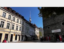 Straßenszene in einer historischen Altstadt mit hellen Gebäuden im klassizistischen Stil links und dem alten Backsteingebäude der Nikolaikirche rechts, an der ein Schild mit „Museum Nikolaikirche“ angebracht ist; im Hintergrund ist der Berliner Fernsehturm sichtbar.