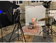 Fotostudio mit professionellem Beleuchtungssetup, das einen Stuhl mit buntem, gemustertem Stoff und passendem Fußhocker auf farbigen Teppichen vor einem weißen Hintergrund zeigt. Umgeben von Kamerastativ, Softboxen und Reflektoren.