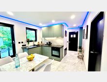 Moderne Küche mit hellgrünen Schränken und dunkler Arbeitsplatte, eingebautem Ofen und Marmor-Rückwand. Im Vordergrund steht ein Esstisch mit Marmoroptik, im Hintergrund sind große Fenster mit Blick ins Freie und eine Eingangstür zu sehen.