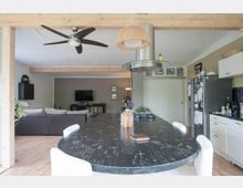 Offener Wohnraum mit Ess- und Küchenbereich, der in einen Wohnbereich übergeht. Im Vordergrund steht ein großer, dunkler Esstisch mit weißen Stühlen, dahinter eine moderne Küche mit weißer Einrichtung und ein Wohnzimmer mit Sofa und Flachbildfernseher. Die Holzdecke ist sichtbar, und ein Deckenventilator sowie Hängelampen ergänzen die Einrichtung.