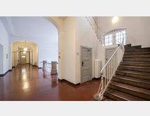 Weitläufiger, heller Flur eines Gebäudes mit glänzendem, rotem Bodenbelag. Rechts führt eine Treppe mit Metallgeländer nach oben, daneben eine graue Tür; links im Hintergrund befinden sich gläserne Durchgangstüren unter einer Rundbogenarchitektur.