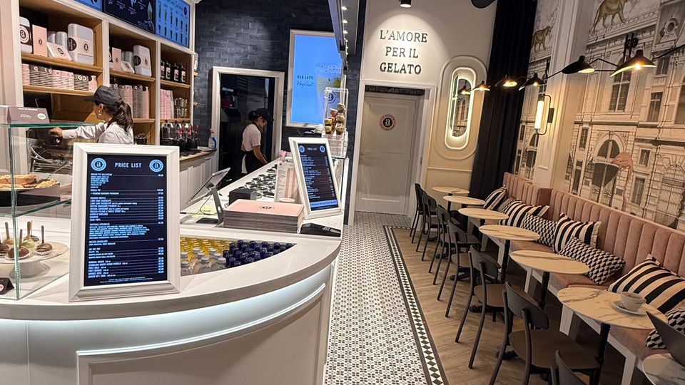 Ein modernes Eiscafé mit geschwungenem, weißem Tresen, auf dem Speisekarten stehen. Links befinden sich Regale mit Produkten, rechts Sitzbereiche mit gepolsterten Bänken, kleinen Holztischen und schwarz-gerahmten Stühlen. Die Wände zeigen kunstvolle Verzierungen und Motive, der Boden ist mit gemusterten Fliesen gestaltet.