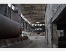 Innenansicht einer verlassenen Fabrikhalle mit hohen Decken, großflächigen Fenstern und sichtbarer alter Bausubstanz. Im Vordergrund ist ein rostiger, zylindrischer Tank zu sehen, umgeben von Beton- und Backsteinwänden.