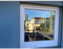 Ansicht aus einem Fenster mit weißem Rahmen, das den Blick auf moderne Gebäude mit flachen Dächern und großen Fenstern freigibt. Im Fenster spiegelt sich ein Teil des Außengeländes mit Bäumen und weiteren Gebäuden.