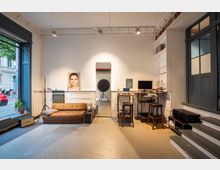 Fotostudio mit schlichtem, modernem Interieur: Ein braunes Sofa steht vor einer weißen Wand mit einem großen Spiegel und einem Porträtfoto. Links ist ein großes Fenster mit Blick auf die Straße, rechts gibt es einen Bereich mit Arbeitsplätzen, Regalen und Computern.