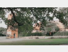 Backsteingebäude mit einem kleinen Turm und roten Dachziegeln, umgeben von Hecken, Bäumen und einer Rasenfläche; im Vordergrund sind Äste eines großen Baumes sichtbar.