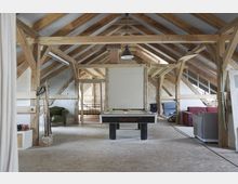 Ein geräumiger Dachboden mit freiliegenden Holzbalken und schrägen Decken, ausgestattet mit einem Billardtisch in der Mitte. Im Hintergrund befinden sich Sitzmöbel, ein Bettgestell und Ziegelwände, die den rustikalen Charakter des Raums unterstreichen.