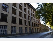 Straßenszene mit einem verlassenen Industriegebäude aus gelbem Backstein, das durch mehrere große, offene Fensteröffnungen und vergitterte Erdgeschossfenster geprägt ist. Auf der rechten Seite säumen Bäume die gepflasterte Straße.