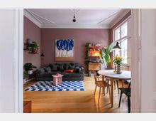 Wohnzimmer mit rosa Wänden, einem dunkelgrauen Sofa, einem blau-weiß gemusterten Teppich und einem runden Esstisch mit Stühlen. Im Hintergrund befinden sich Pflanzen, ein Regal mit Büchern und Dekoration sowie ein großes Fenster, das Tageslicht hereinlässt.