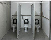 Öffentlicher Toilettenraum mit drei nebeneinander platzierten Toiletten, die durch einfache Trennwände ohne Türen voneinander getrennt sind. Die Wände sind weiß, der Boden ist aus grauem Beton, und es sind sichtbare Rohrleitungen an den Wänden angebracht.