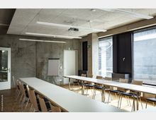 Konferenzraum mit langen weißen Tischen und modernen Holzstühlen auf Parkettboden. Die Wände sind aus Sichtbeton, es gibt eine große Fensterfront mit schwarzen Jalousien, und ein Flipchart steht in der Ecke.