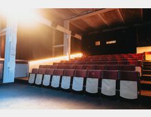 Ein kleiner Theater- oder Kinosaal mit mehreren Reihen roter Polstersitze und klappbaren Holzsitzen. Die Beleuchtung ist warm, und im Hintergrund sind eine schwarze Wand mit zwei kleinen Fenstern sowie ein freiliegendes Decken- und Tragwerksdesign sichtbar.