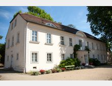 Zweistöckiges, hell verputztes Gebäude mit Satteldach und weißen Fensterrahmen, umgeben von Pflanzen und Blumenbeeten in einem Hof mit Kiesboden. Eine kleine Treppe führt zu einer zentralen Tür mit einem Balkon darüber.