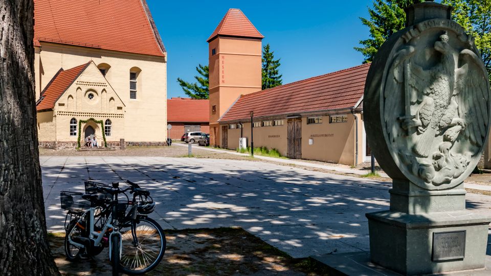 Großer gepflasterter Platz mit historischen Gebäuden mit roten Ziegeldächern, darunter eine Kirche mit Rundbogenportal und ein turmartiger Bau. Im Vordergrund stehen ein steinernes Denkmal mit Relief eines Adlers sowie Fahrräder neben einem großen Baum; im Hintergrund sind Bäume und weitere Gebäude zu sehen.